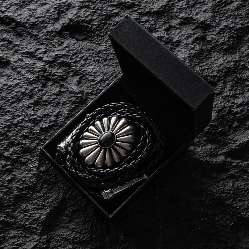 May include: A black bolo tie with a silver-colored floral design and a black stone center, presented in a black box. The braided leather cord is coiled within the box, with the silver-toned tips visible.