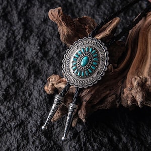 Pode incluir: Gravata bolo prateada com uma pedra central turquesa e detalhes a condizer. O pendente oval é rodeado por pedras turquesas mais pequenas e uma borda prateada decorativa. O cordão de couro preto tem pontas prateadas. Joalharia de estilo Western.