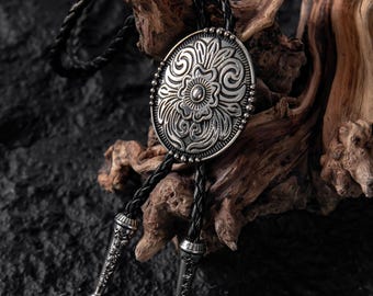 May include: A bolo tie featuring an oval silver-toned metal centerpiece with an embossed floral and scrollwork pattern, bordered by small metal beads. The centerpiece is attached to a black braided leather cord with two matching silver-toned metal tips.