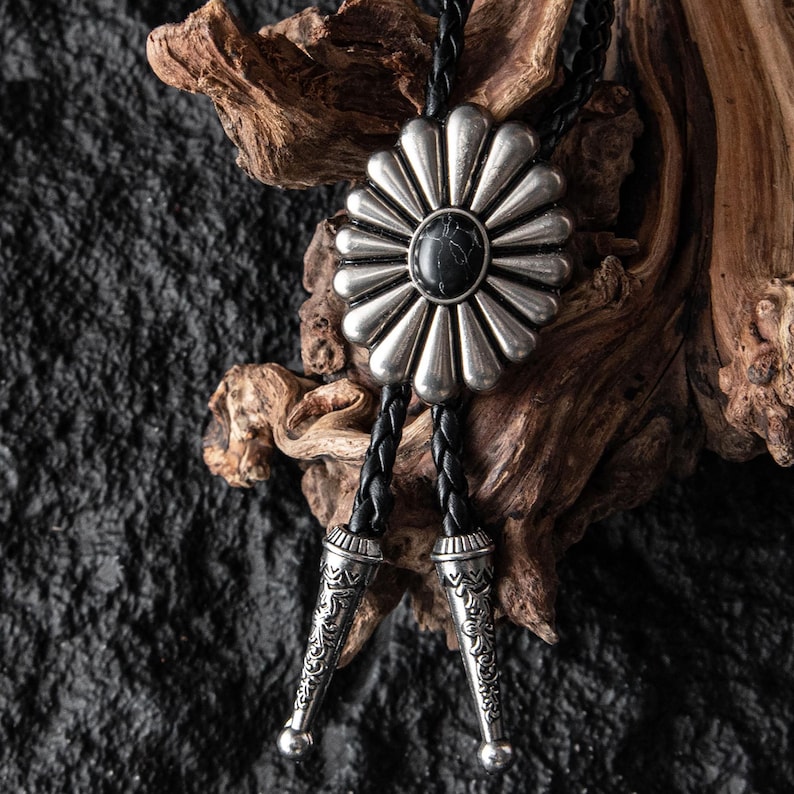 May include: A silver bolo tie with a large, flower-shaped pendant featuring a black stone center. The pendant is connected to a black braided leather cord with decorative silver tips. The bolo tie is a Western-style accessory.