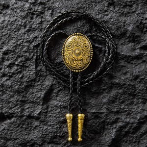 May include: A bolo tie with a gold-colored oval pendant featuring an ornate floral design. The pendant is surrounded by small, gold-colored beads. The black braided leather cord has gold-colored tips. The tie is set against a dark, textured background.