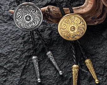 May include: Two bolo ties with circular, floral-patterned pendants. One is silver-toned, the other gold-toned. Both have black braided cords and decorative tips. The ties rest on a dark, textured surface with a piece of wood.