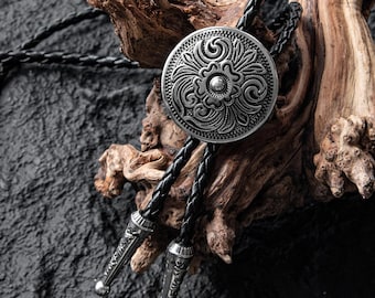 May include: A silver bolo tie with a circular, floral-patterned pendant. The pendant is attached to two black braided cords with decorative silver tips. The bolo tie is displayed against a rustic wooden background.