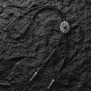 May include: A silver bolo tie with a black braided cord. The bolo tie features a flower-shaped pendant with a black center stone. The cord ends have decorative silver tips. The tie is set against a textured black background.