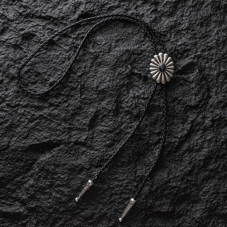 May include: A silver bolo tie with a black braided cord. The bolo tie features a flower-shaped pendant with a black center stone. The cord ends have decorative silver tips. The tie is set against a textured black background.
