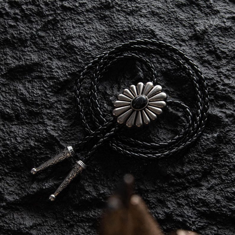 May include: A black leather bolo tie with a silver-toned daisy-shaped pendant featuring a black stone center. The braided cord is looped, and the tips are silver with etched designs. The bolo tie rests on a textured black surface.