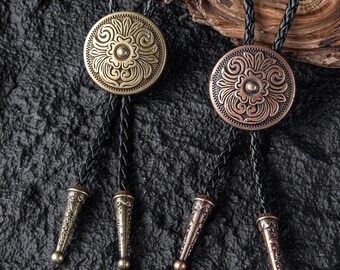 May include: Two bolo ties with circular, embossed metal pendants featuring floral designs. The pendants are in antique gold and copper tones, with black braided cords and decorative tips. The ties are displayed against a textured, dark background.