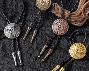 May include: Several bolo ties with round, ornate metal pendants and braided black cords. The pendants are in silver, bronze, copper, and gold tones, each with a detailed floral design. The tips of the ties are also metallic.