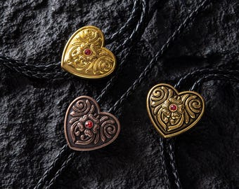 May include: Three heart-shaped bolo ties with intricate floral designs and a central red gem. The bolo tie pendants are in gold and bronze tones, attached to black braided cords. The background is a textured black surface.