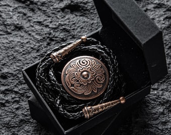 May include: A bolo tie with a copper-colored, circular pendant featuring an embossed floral design. The pendant is connected to a braided black leather cord, presented in a black box. The tips of the cord are also copper-colored.