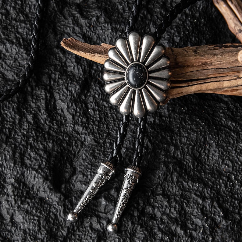 May include: A silver bolo tie with a black stone center and a floral design. The tie features a black braided cord and decorative silver tips. The bolo tie is a western-style accessory.