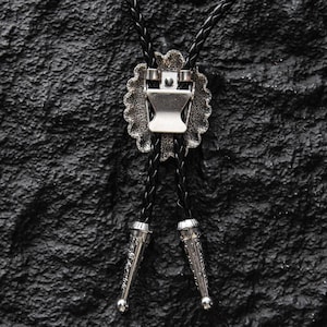 May include: A close-up of a bolo tie against a textured black surface. The tie features a silver-colored, scalloped-edge pendant with a central clasp. Two braided black cords extend downwards, each ending in a decorative silver tip.