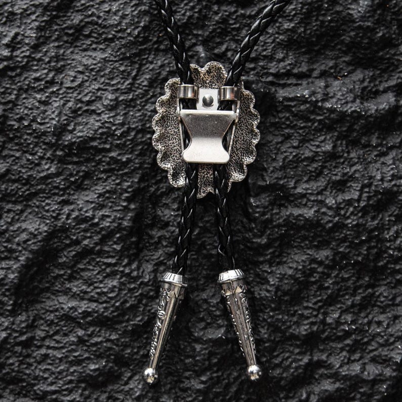 May include: A close-up of a bolo tie against a textured black surface. The tie features a silver-colored, scalloped-edge pendant with a central clasp. Two braided black cords extend downwards, each ending in a decorative silver tip.