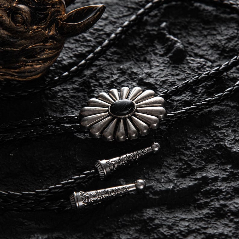 May include: A silver bolo tie with a flower-shaped pendant featuring a black stone center. The pendant is connected to a black braided leather cord with decorative silver tips. The bolo tie rests on a dark, textured surface.