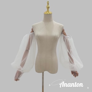 Puede incluir: Mangas blancas con hombros descubiertos y tejido vaporoso. Las mangas están sujetas a un maniquí, mostrando el diseño. El texto "Ananton" es visible en la esquina inferior derecha. Las mangas tienen una apariencia delicada y aireada.