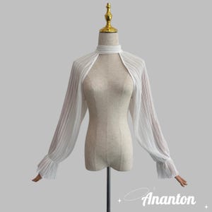 Puede incluir: Un bolero blanco y transparente con mangas largas plisadas y un cuello alto y abierto. Las mangas terminan en puños con volantes. El bolero se muestra en un maniquí. La marca "Ananton" está impresa en una fuente cursiva en la parte inferior derecha.