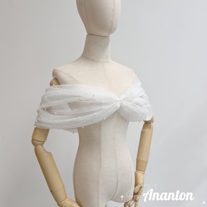 May include: A white, off-the-shoulder bridal wrap made of sheer fabric, adorned with small, scattered pearls. The wrap is draped over a mannequin, showcasing its design and texture. The wrap is tied in a bow at the center.