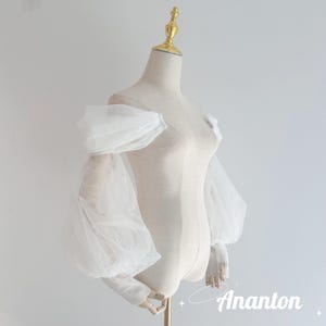Puede incluir: Una blusa blanca con hombros descubiertos y mangas abullonadas transparentes. La prenda se exhibe en un maniquí con un adorno dorado para la cabeza. Las mangas están recogidas en la muñeca y el hombro, creando un efecto voluminoso. La marca "Ananton" es visible.