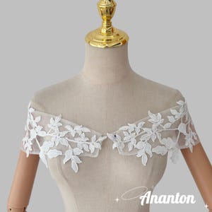 May include: White lace bridal shoulder wrap with a floral leaf design. The delicate lace overlay is displayed on a mannequin. The wrap is designed to be worn over the shoulders. The brand name "Ananton" is visible.