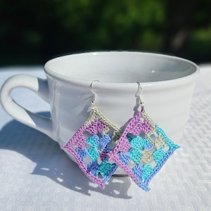 May include: A pair of crocheted earrings with a square shape. The earrings are made with a combination of light purple, light blue, and light green yarn. The earrings are hanging from silver hooks.