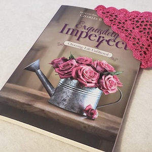 May include: A book titled "Exquisitely Imperfect: Choosing Life Unfiltered" by Charles R. Swindoll. The book cover features a vintage watering can filled with pink roses and a pink crocheted bookmark.
