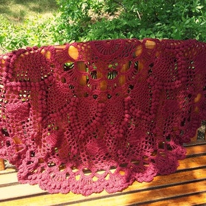 May include: A burgundy-colored, handmade crochet shawl with intricate openwork patterns. The shawl is draped over a wooden bench, showcasing its detailed floral and geometric designs. The yarn appears to be a soft, natural fiber.