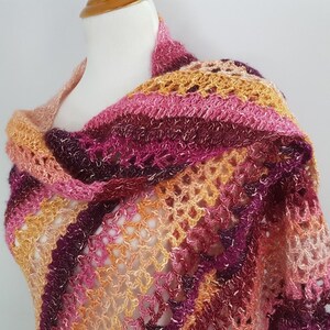 May include: A handmade, open-weave shawl in shades of pink, orange, and burgundy. The shawl is draped over a mannequin, showcasing its intricate crochet pattern and color gradient. The design features a mix of solid and open sections.