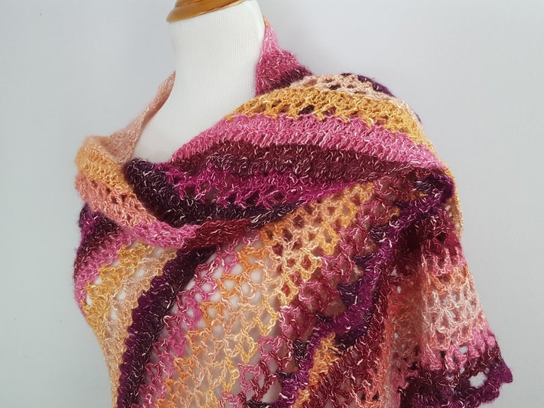 May include: A handmade, open-weave shawl in shades of pink, orange, and burgundy. The shawl is draped over a mannequin, showcasing its intricate crochet pattern and color gradient. The design features a mix of solid and open sections.