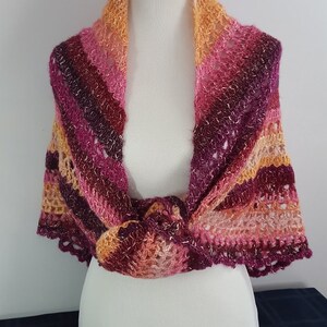 May include: A handmade, open-weave shawl in shades of pink, burgundy, and orange. The shawl is draped over a mannequin, showcasing its intricate crochet pattern and scalloped edges. The color gradient creates a warm, inviting aesthetic.