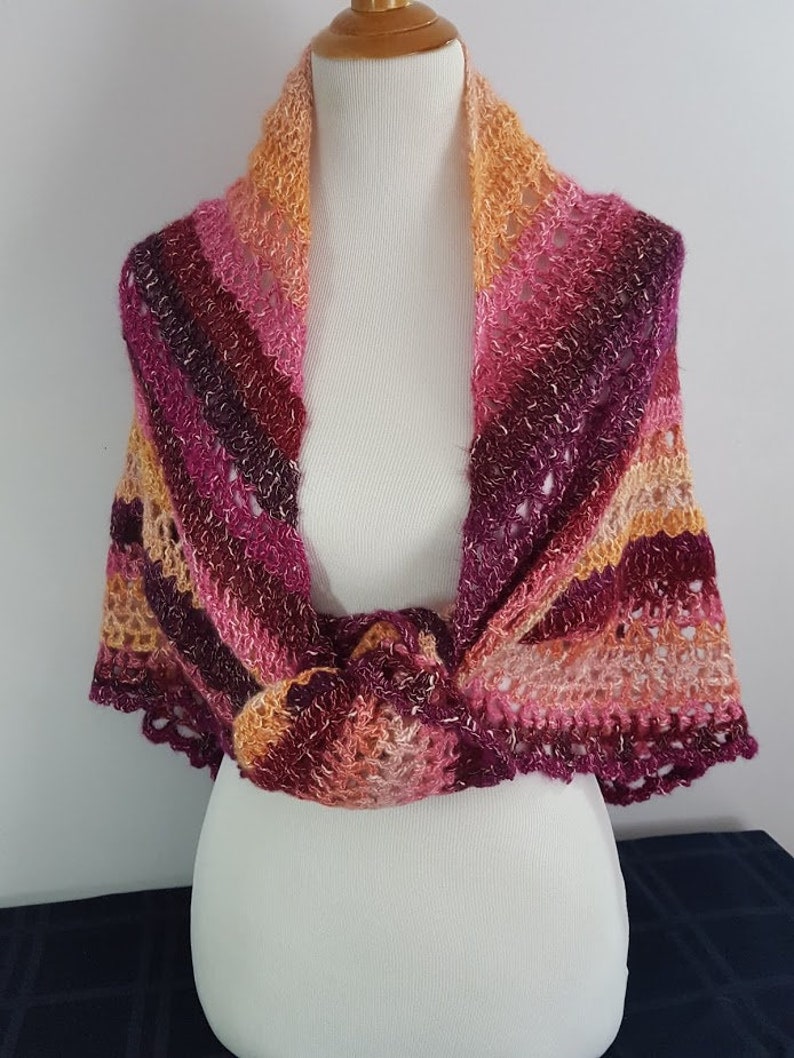 May include: A handmade, open-weave shawl in shades of pink, burgundy, and orange. The shawl is draped over a mannequin, showcasing its intricate crochet pattern and scalloped edges. The color gradient creates a warm, inviting aesthetic.