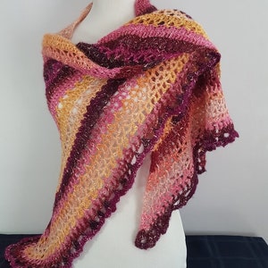 May include: A handmade, open-weave shawl in shades of pink, orange, and burgundy. The shawl is draped over a mannequin, showcasing its intricate crochet design and scalloped edges. The color gradient creates a warm, inviting aesthetic.