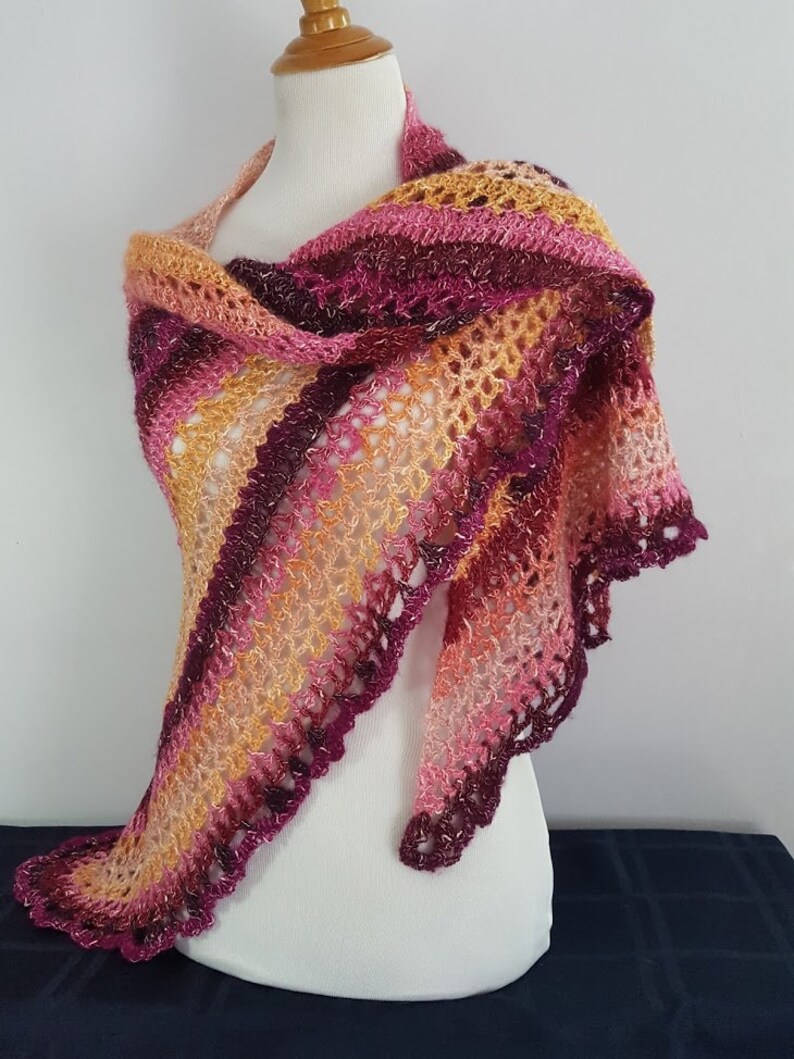 May include: A handmade, open-weave shawl in shades of pink, orange, and burgundy. The shawl is draped over a mannequin, showcasing its intricate crochet design and scalloped edges. The color gradient creates a warm, inviting aesthetic.