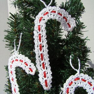 May include: Three white crocheted candy cane ornaments with red ribbon accents hanging on a green artificial Christmas tree.