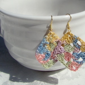 May include: A pair of handmade crochet earrings in a diamond shape. The earrings feature a colorful patchwork design with pastel shades of pink, blue, yellow, and green. The earrings are suspended from gold-colored ear wires.