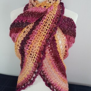 May include: A handmade, open-weave scarf in shades of pink, burgundy, and gold, draped over a mannequin. The scarf features a scalloped edge and a striped pattern, showcasing a blend of warm colors.
