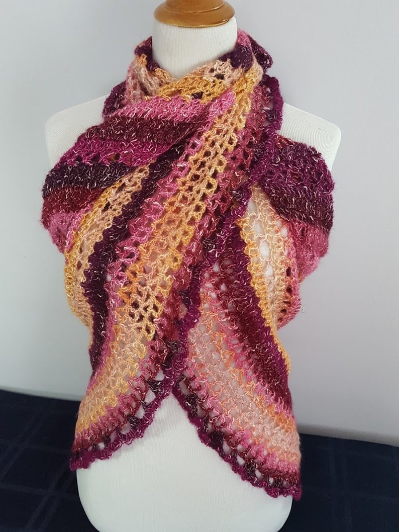 May include: A handmade, open-weave scarf in shades of pink, burgundy, and gold, draped over a mannequin. The scarf features a scalloped edge and a striped pattern, showcasing a blend of warm colors.