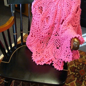 May include: A pink crocheted blanket draped over a black wooden chair. The chair has a high back and a curved seat. The blanket is made of delicate, openwork stitches.