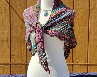 May include: A handmade crochet shawl in shades of purple, teal, and green is draped over a mannequin. The shawl has an open, lacy design and is tied in the front. A silver necklace with a square pendant is also displayed.