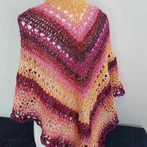 May include: A handmade shawl in a vibrant array of colors, including shades of pink, orange, and burgundy. The shawl features a delicate, openwork crochet design with a scalloped edge. The colors transition in stripes, creating a visually appealing accessory.