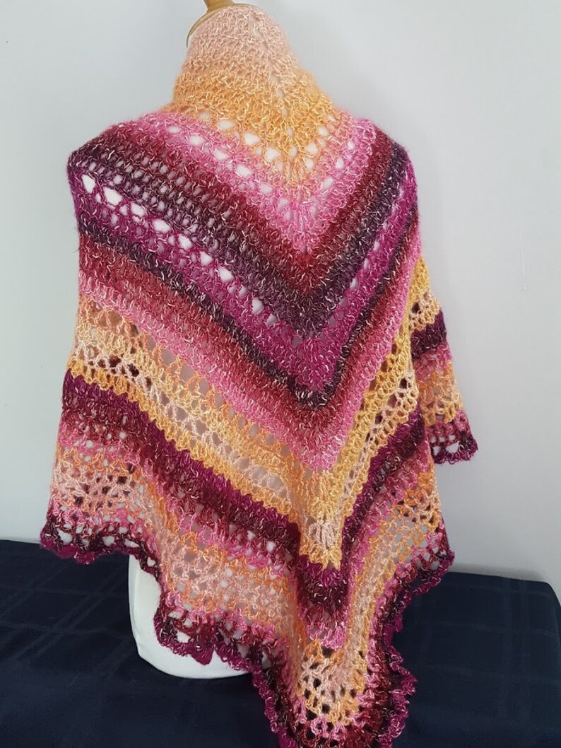 May include: A handmade shawl in a vibrant array of colors, including shades of pink, orange, and burgundy. The shawl features a delicate, openwork crochet design with a scalloped edge. The colors transition in stripes, creating a visually appealing accessory.