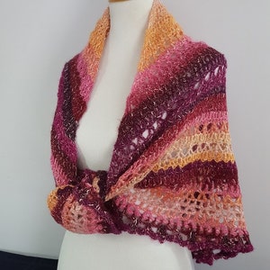 May include: A handmade, colorful crochet shawl in shades of orange, pink, and purple. The shawl is draped over a mannequin, showcasing its open weave and knotted design. The edges have a scalloped trim.
