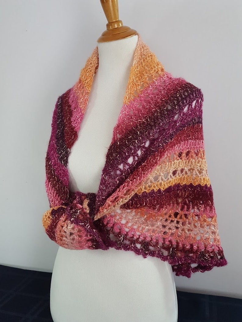 May include: A handmade, colorful crochet shawl in shades of orange, pink, and purple. The shawl is draped over a mannequin, showcasing its open weave and knotted design. The edges have a scalloped trim.