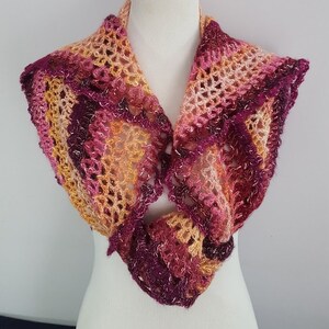 May include: A handmade crochet shawl in shades of pink, purple, and gold. The openwork design creates a delicate, lacy texture. The shawl is displayed on a white mannequin, showcasing its shape and drape. The color palette is warm and inviting.