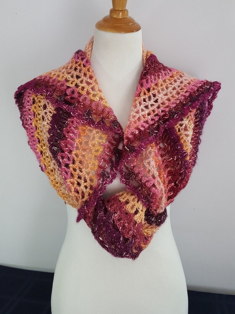 May include: A handmade crochet shawl in shades of pink, purple, and gold. The openwork design creates a delicate, lacy texture. The shawl is displayed on a white mannequin, showcasing its shape and drape. The color palette is warm and inviting.