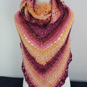 May include: A handmade, triangular shawl in shades of pink, orange, and burgundy, displayed on a mannequin. The shawl features a lacy, openwork design with a scalloped edge. The color gradient transitions from light to dark.