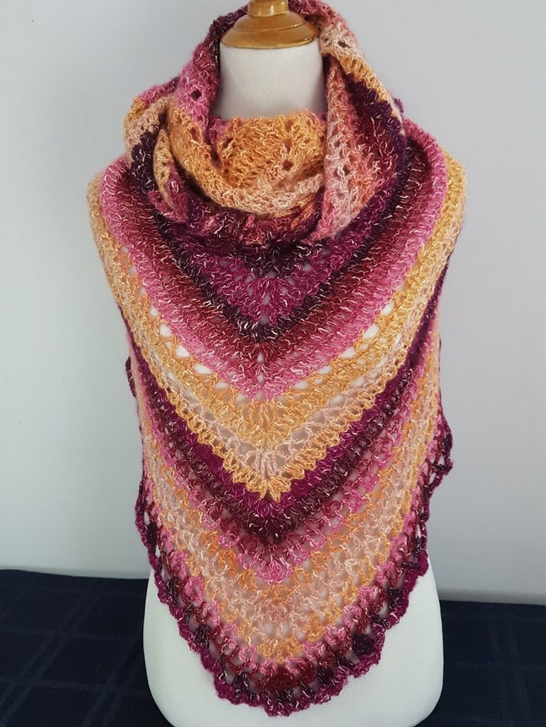May include: A handmade, triangular shawl in shades of pink, orange, and burgundy, displayed on a mannequin. The shawl features a lacy, openwork design with a scalloped edge. The color gradient transitions from light to dark.