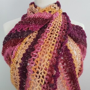 May include: A handmade, open-weave shawl in shades of pink, purple, and orange. The scarf is draped over a mannequin, showcasing its intricate crochet pattern and color gradient. The yarn appears soft and textured.