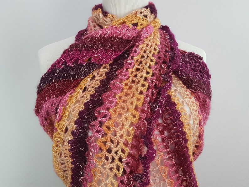 May include: A handmade, open-weave shawl in shades of pink, purple, and orange. The scarf is draped over a mannequin, showcasing its intricate crochet pattern and color gradient. The yarn appears soft and textured.