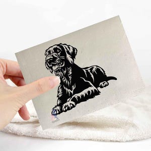 May include: A black and white illustration of a Schnauzer dog in a relaxed pose. The dog is lying down, with its front paws extended. The image is on a light beige square, held by a hand, resting on a white towel.