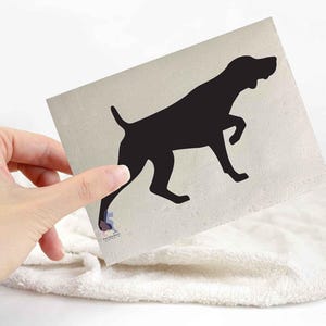 May include: A beige square card with a black silhouette of a dog in a pointing pose. The dog is facing right with one leg raised. The card is held over a white, fluffy surface.